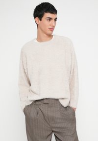 Pulover bej pufos, cu gât rotund, croială lejeră, textură moale, asortat cu pantaloni maro cu dungi subțiri. Design simplu, cu umeri căzuți.