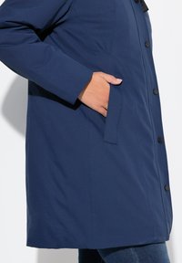 Cappotto impermeabile blu navy con una texture liscia, dotato di una tasca laterale e bottoni neri lungo la parte anteriore, progettato per una vestibilità comoda.