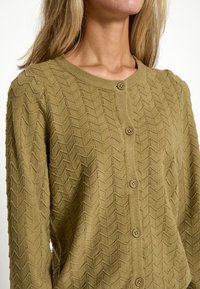 Khaki zielony sweter z dzianiny, z fakturowanym wzorem w zygzaki, pięcioma zapięciami na guziki oraz okrągłym dekoltem. Miękki, lekki materiał.