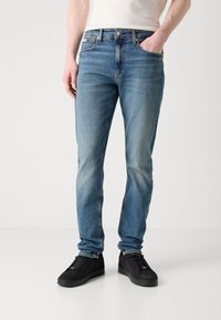 Light blue denim jeans with a slim fit, five-pocket design, slight fading, and visible stitching details. Paired with black sneakers.