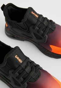Une paire de baskets noires et orange avec une étiquette "GAMER" sur les languettes rembourrées et de grosses lacets noirs, sur un fond gris clair.