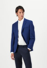 Marineblå blazer med tilpasset fit, notch-lapper, en brystlomme og to-knaps lukking, båret over en hvit skjorte og mørke bukser.