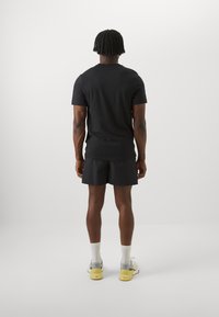 T-shirt nera a maniche corte e colletto rotondo abbinata a pantaloni corti neri con texture. Calze bianche e scarpe da ginnastica grigie/gialle completano il look.
