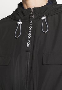 Giacca nera con chiusura frontale con cerniera, cappuccio e due tasche anteriori. Dotata di coulisse con punte metalliche e marchio sulla cerniera.