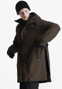 Parka impermeabile marrone con cappuccio, tasche con cerniera e una texture liscia. Il modello indossa guanti neri e un berretto, con una vestibilità rilassata.