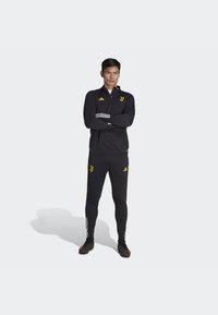 Schwarzer Sport-Trainingsanzug mit gelbem Logo und Akzenten. Zip-Up-Oberteil mit drei Streifen an den Ärmeln; eng anliegende Hose mit Logo auf den Taschen.
