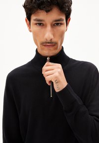 Zwarte zip-neck trui van gladde stof. Heeft een hoge kraag en zilveren ritsdetails. Model heeft een licht getatoeëerde hand.