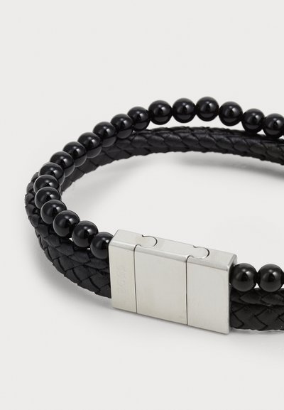 Bracelet en cuir tissé noir avec des perles noires lisses, fermoir métallique avec logo gravé et une disposition de perles circulaires.