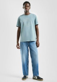 Jasnoniebieski t-shirt z krótkim rękawem z okrągłym logo, w zestawie z luźnymi, jasnoniebieskimi dżinsami i czarnymi trampkami.