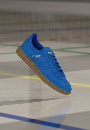 Blauer Adidas Spezial Sneaker mit Gummisohle, der über einem Sporthallenboden schwebt, der mit Linien eines Sportplatzes markiert ist.
