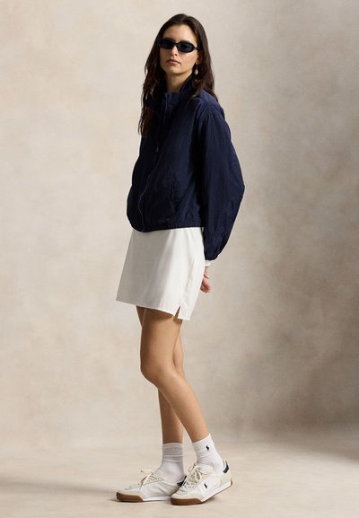 Marineblå bomberjakke med glidelås, hvit plissert skjørt, hvite joggesko med marineblå detaljer, og ribbestrømper. Modellen står foran en nøytral bakgrunn.