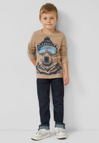 Beige shirt met lange mouwen met een beerafbeelding met blauwe zonnebril, gecombineerd met donkerblauwe spijkerbroek en witte sneakers met grijze accenten.