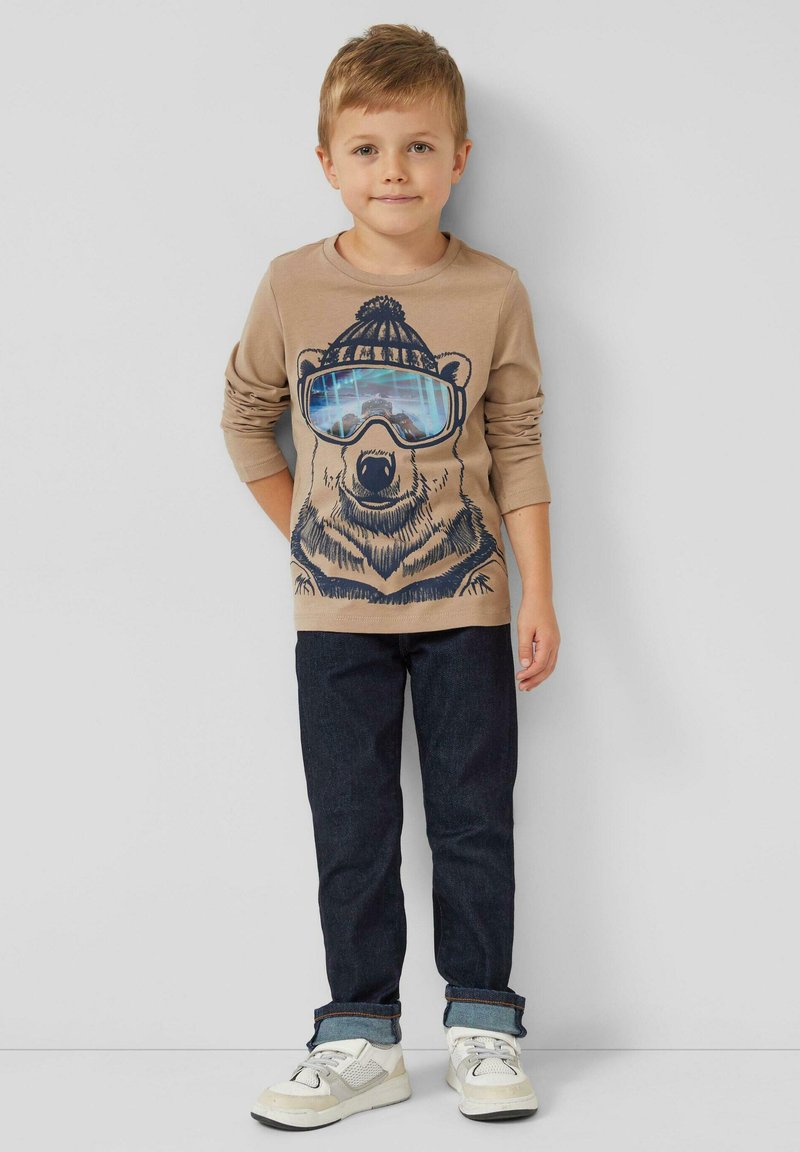 Beige shirt met lange mouwen met een beerafbeelding met blauwe zonnebril, gecombineerd met donkerblauwe spijkerbroek en witte sneakers met grijze accenten.