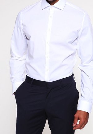 Mand iført en tætsiddende hvid skjorte med knapper, stoppet ned i sorte bukser, stående med den ene hånd i lommen.