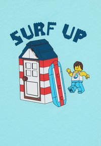 Lego figuur vaba aja riietuses triibulise rannaonni ja surfilaudade lähedal. Taust on helesinine, kujunduse kohal on tekst "SURF UP".
