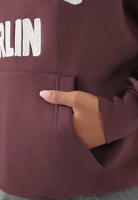 Mano con esmalte de uñas rosa descansando dentro del bolsillo de una sudadera burdeos con el texto blanco "BERLÍN" encima del bolsillo.