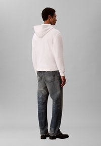 Vīrietis stāv ar muguru pret mums, tērpies baltā sporta kapucē, izbalējušās zilās džinsu biksēs un melnās kurpēs, fona krāsa viendabīgi pelēka.