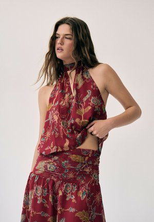 Jeune femme aux longs cheveux bruns portant un haut dos nu à fleurs rouge et une jupe assortie, posant avec une main près de sa taille.