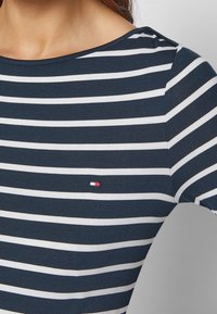 Mujer vistiendo una camiseta de rayas horizontales azul marino y blanca con un pequeño logo rectangular rojo, blanco y azul en el pecho.