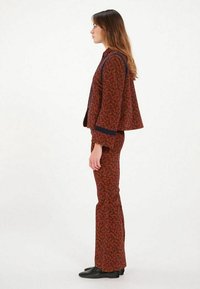 Costume rayé rouge, comprenant une veste ajustée avec des manches évasées et un pantalon évasé assorti. Des accents en bordure sombre soulignent le design.