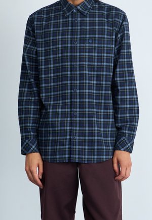 Mężczyzna w niebiesko-czarnej kratowanej koszuli na guziki z długim rękawem i kieszenią na piersi, w ciemnobrązowych spodniach, stoi na tle jednolitego tła.