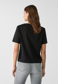 Sort bomulds t-shirt med korte ærmer og rund hals, der har en afslappet pasform. Grå denim jeans med baglommer er synlige.