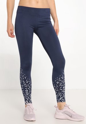 Leggings bleu marine avec une texture lisse et taille haute, ornés d'un motif de feuilles blanches au niveau des chevilles. Associés à des chaussures de sport roses claires.