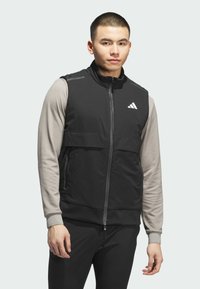 Crni atletski prsluk s patentnim zatvaračem sprijeda, sive duge rukave i Adidas logo. Ima glatku tkaninu, visoki ovratnik i bočne džepove.