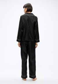 Kobieta stojąca boso tyłem, ubrana w czarne satynowe piżamy z delikatnym nadrukiem w cętki i jasnoróżowym wykończeniem na mankietach i kołnierzu.