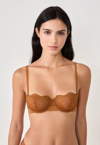 Sostén de encaje bronce con bordes ondulados, que presenta tirantes delgados ajustables y una banda inferior suave; se aprecian patrones intrincados en el encaje.