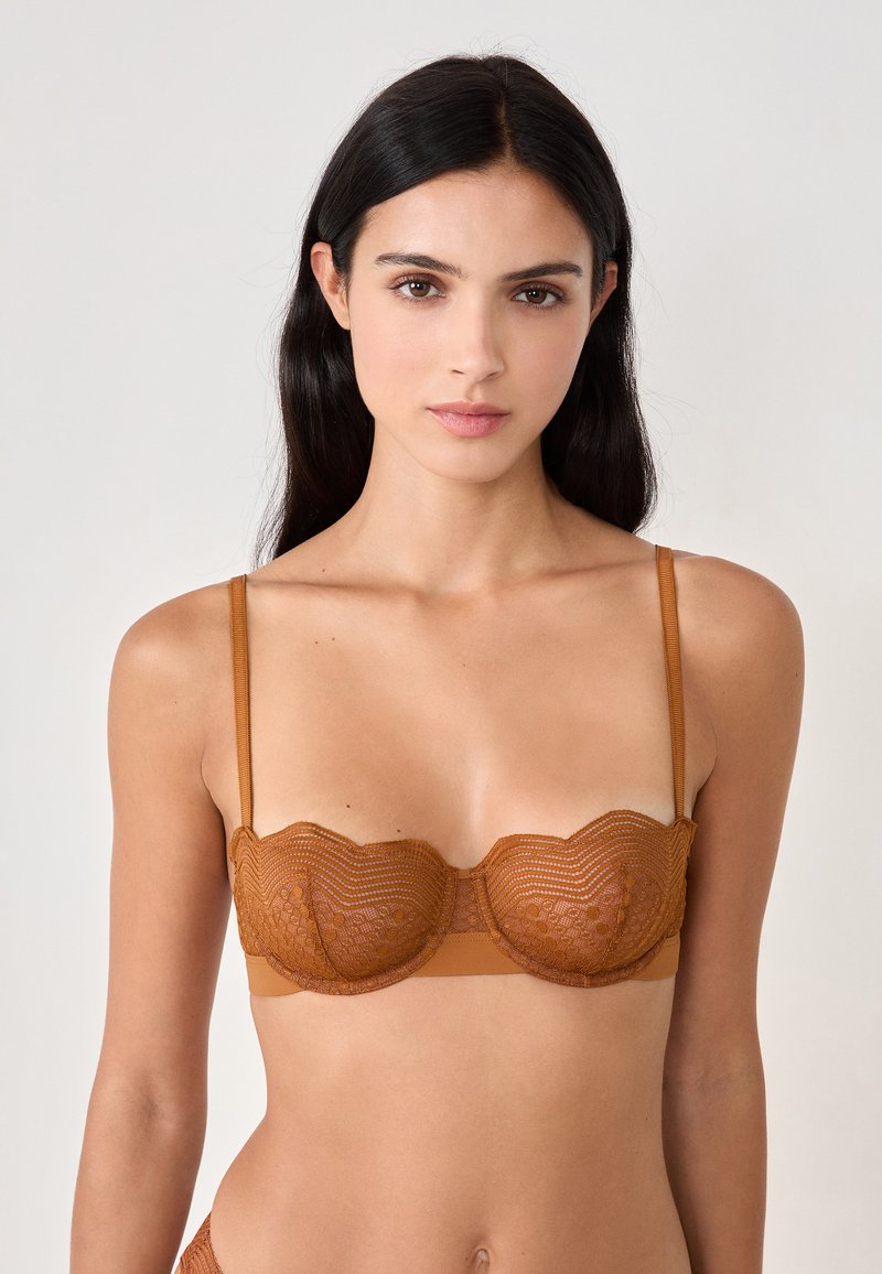 Sostén de encaje bronce con bordes ondulados, que presenta tirantes delgados ajustables y una banda inferior suave; se aprecian patrones intrincados en el encaje.