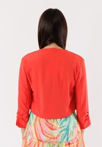 Giacca rossa corta con maniche a tre quarti e texture liscia, abbinata a una gonna floreale a strati e dai colori vivaci.