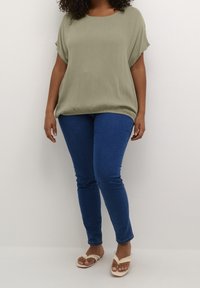 Mujer vestida con una blusa corta de manga corta suelta de color verde claro, jeans ajustados azules y chanclas beige, de pie contra un fondo blanco liso.
