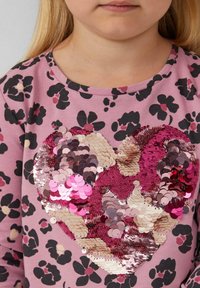 Rosa Langarmshirt mit einem schwarz-lila Blumendruck; verfügt über ein großes Herz aus mehrfarbigen Pailletten in rosa und beige.