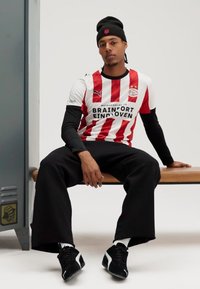 Baltai ir raudonai dryžuotas futbolo marškinėliai su juodais akcentais, juodos kelnės ir juodi sportbačiai. Modelis sėdi ant medinės suoliuko neutraliame fone.