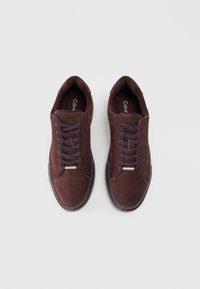 Zapatillas de ante marrón con puntera redonda, cordones planos y detalle metálico en la lengüeta. Plantilla acolchada y suela de goma texturizada.