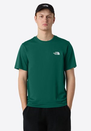 Jeune homme portant un t-shirt The North Face vert foncé et une casquette noire, debout avec les mains dans les poches devant un fond uni.