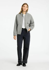 Grijze zip-up jack met hoge kraag, voorzakken en geribbelde mouwen, gedragen met donkere, wijde jeans en zwarte schoenen. Effen witte shirt eronder.