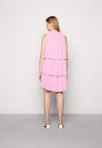 Vestido cor-de-rosa em camadas com um design sem mangas, apresentando um decote com babados e padrão de bolinhas. A modelo está a usar sandálias brancas de salto bloco.