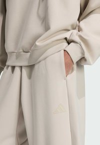 Béžová mikina s kapucňou a tepláky v mäkkej látke. Obsahuje rebrované manžety a akcent s logom Adidas na teplákoch. Uvoľnený a pohodlný strih.