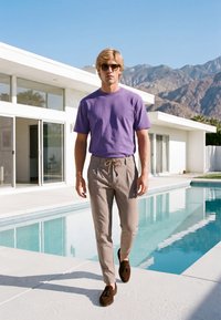Homme portant des lunettes de soleil, un t-shirt violet et un pantalon beige, marchant près d'une piscine devant une maison blanche moderne avec des montagnes en arrière-plan.