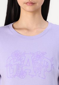 Valkoinen puuvillapaita, jossa on vaaleansininen kukkakuosi, joka on kytketty 'GAP' -logoja vähän tummemmalla violettisävyllä. Pyöreä kaula-aukko.