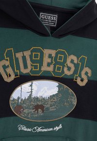 Felpa con cappuccio verde scuro e nera, con grande logo "GUESS" ricamato, toppa con orso paesaggistico e testo "Classic American style" in bianco.