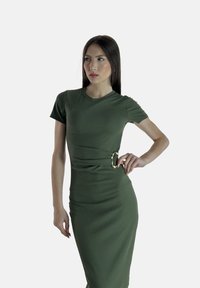 Olivgrünes figurbetontes Kleid mit gerippter Textur, kurzen Ärmeln und einem goldenen ovalen Akzent an der Taille, der einen drapierten Effekt erzeugt.