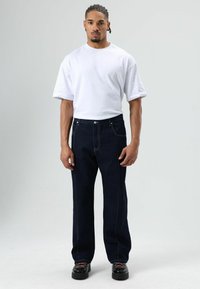 Witte shirt met korte mouwen, donkerblauwe jeans met contrasterende stiksels en zwarte schoenen met kleurrijke veters. Model staat rechtop tegen een effen achtergrond.