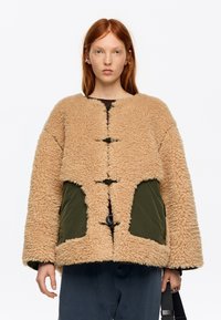 Persona pelirroja que lleva una chaqueta beige y esponjosa con bolsillos verdes y botones de palanca, sosteniendo una correa negra, sobre un fondo blanco liso.