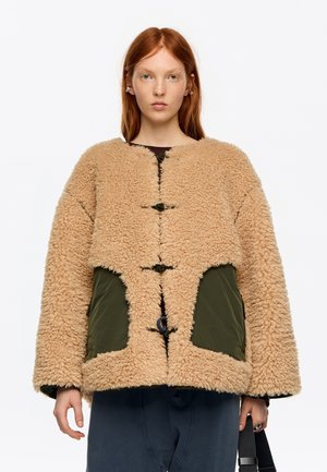 Persona pelirroja que lleva una chaqueta beige y esponjosa con bolsillos verdes y botones de palanca, sosteniendo una correa negra, sobre un fondo blanco liso.