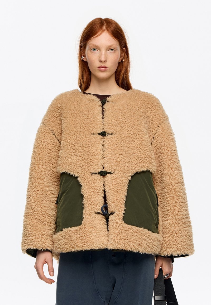 Persona pelirroja que lleva una chaqueta beige y esponjosa con bolsillos verdes y botones de palanca, sosteniendo una correa negra, sobre un fondo blanco liso.