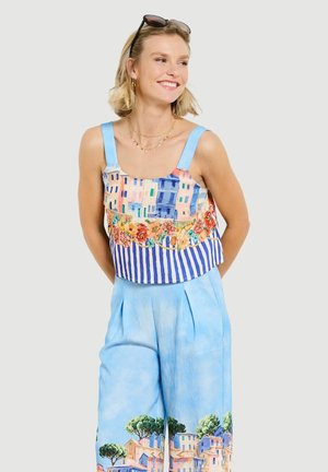 Jeune femme souriante, portant un pantalon bleu à jambes larges et un haut sans manches avec des motifs colorés de bâtiments et de fleurs.