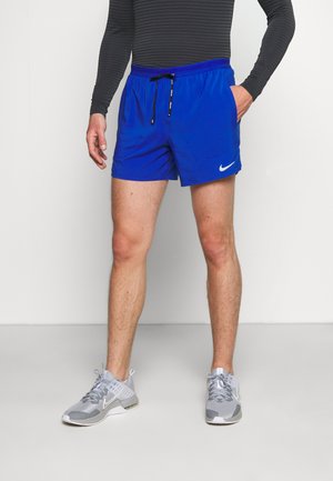 Homme portant un short Nike bleu vif et des baskets Nike grises, debout avec une main dans la poche devant un fond uni.
